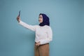 A portrait of a happy Asian Muslim woman wearing a white shirt and hijab, holding her phone, isolated by blue background Royalty Free Stock Photo