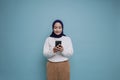 A portrait of a happy Asian Muslim woman wearing a white shirt and hijab, holding her phone, isolated by blue background Royalty Free Stock Photo