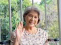 Portrait of happy Asian senior woman grey hair sitting on wheelchair, waving greeting, smiling and looking at camera.