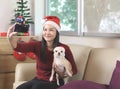 Happy Asian woman wearing Christmas Santa hat sitting  in living room with Christmas decoration, taking selfie photo with her Royalty Free Stock Photo