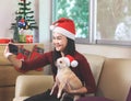 Asian woman wearing Christmas Santa hat sitting  in living room with Christmas decoration, taking selfie photo with her chihuahua Royalty Free Stock Photo