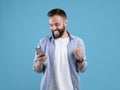 Portrait of happy bearded young man using smartphone, making winner gesture over blue studio background Royalty Free Stock Photo