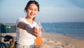 Portrait of happy beautiful Asian woman sitting on a picnic chair holding orange soft drink bottle and drinking on beach in summer