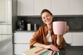 Portrait of happy, beautiful brunette woman in kitchen, student taking lunch break for cup of tea, raising mug, giving