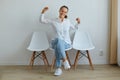 Portrait of happy beautiful female wearing white shirt and jeans raised arms, stretching her hands while sitting on chair against Royalty Free Stock Photo