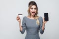 Portrait of happy beautiful modern young girl in grey dress standing, holding cell phone and credit card with open mouth, looking Royalty Free Stock Photo