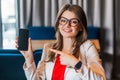 Portrait of happy beautiful stylish young woman in glasses sitting, showing and pointing at mobile smart phone screen and looking Royalty Free Stock Photo
