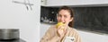 Portrait of happy, beautiful woman smiling, eating an apple in the kitchen, sitting at home in bathrobe, holding Royalty Free Stock Photo