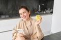Portrait of happy, beautiful woman smiling, eating an apple in the kitchen, sitting at home in bathrobe, holding Royalty Free Stock Photo