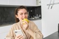 Portrait of happy, beautiful woman smiling, eating an apple in the kitchen, sitting at home in bathrobe, holding Royalty Free Stock Photo