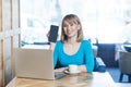 Portrait of happy beautiful young blonde woman in blue t-shirt, sitting with laptop, holding and showing mobile display and Royalty Free Stock Photo