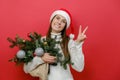 Portrait of happy beautiful young woman in Santa Claus hat holding bouquet of spruce branches show victory sign, posing isolated Royalty Free Stock Photo