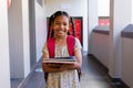 Portrait of happy biracial schoolgirl with books and school bag at elementary school corridor Royalty Free Stock Photo