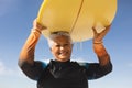 Portrait of happy biracial senior woman carrying yellow surfboard overhead against blue sky Royalty Free Stock Photo