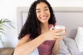 Portrait of happy biracial woman holding cup of coffee sitting on bed at sunny home Royalty Free Stock Photo