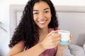 Portrait of happy biracial woman holding cup of coffee sitting on bed at sunny home Royalty Free Stock Photo