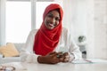 Portrait of happy black muslim businesswoman in hijab using cellphone while sitting at desk and smiling Royalty Free Stock Photo