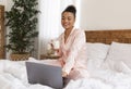 Portrait of happy black woman sitting on bed, using modern laptop computer and drinking coffee, enjoying morning Royalty Free Stock Photo