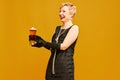 Portrait of happy blond mature woman in retro black dress holding beer glass and smiling over yellow studio background Royalty Free Stock Photo