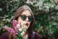 Portrait of happy boho girl in sunglasses smiling with bouquet of wildflowers in sunny garden. Stylish hipster carefree girl