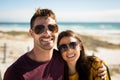 Portrait of happy caucasian couple sitting on beach by the sea embracing wearing sunglasses smiling Royalty Free Stock Photo