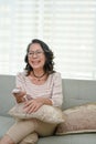 Happy 60s retired Asian woman wearing glasses, sitting on couch, enjoys watching TV Royalty Free Stock Photo