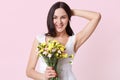 Portrait of a happy cheerful young woman in summer white dress holding colourful flowers in one hand, fixing her hair, looking Royalty Free Stock Photo