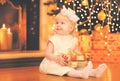 Portrait happy child little girl sitting with gift box looking away near christmas tree with garland lights and fireplace at Royalty Free Stock Photo