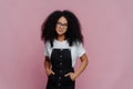 Portrait of happy curly African American woman keeps hands in pockets of sarafan, wears transparent glasses, meets with friends Royalty Free Stock Photo