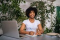 Portrait of happy curly African-American young woman wearing headphones holding pen in hand sitting at desk with laptop Royalty Free Stock Photo