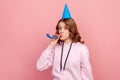 Portrait of happy curly haired teenage girl in hoodie with birthday hat blowing party horn, having fun at holiday celebration Royalty Free Stock Photo
