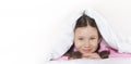 Portrait of a happy cute girl lying on white bed linen under a blanket. Copy space for text Royalty Free Stock Photo