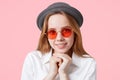 Portrait of happy delighted female model in round red sunglasses and elegant hat, keeps hands under chin, smiles broadly, isolated