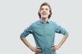 Portrait of happy dreamy handsome long haired blonde young man in blue casual shirt standing with hands on waists and looking away Royalty Free Stock Photo