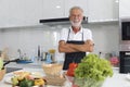 Portrait of happy elderly man with apron standing with arms crosse at kitchen with colorful fresh vegetables, fruits,ingredients.