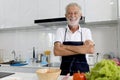 Portrait of happy elderly man with apron standing with arms crosse at kitchen with colorful fresh vegetables, fruits,ingredients.