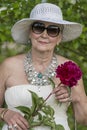 Portrait of a happy elderly woman 65 - 70 years old in a white straw hat with red peony flower, closeup Royalty Free Stock Photo