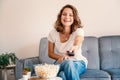 Portrait of happy emotional beautiful young girl woman in jeans couch at home eating popcorn and watching TV in remote control in Royalty Free Stock Photo