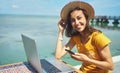 Portrait happy freelancer woman in hat smiling to camera, holding smartphone, working on laptop computer by seashore Royalty Free Stock Photo