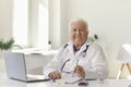 Friendly senior doctor sitting at desk with laptop in modern hospital office and looking at camera Royalty Free Stock Photo