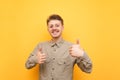 Portrait of happy geek in glasses and beige shirt stands on yellow background, shows thumbs up and looks into camera with smile on Royalty Free Stock Photo