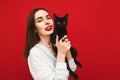 Portrait of happy girl with black cat in hands on red background, smiling and posing at camera. Cute positive lady in white shirt Royalty Free Stock Photo