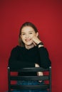 Portrait of happy girl in black sweater and clock on her hand sits on a chair and smiles isolated on red background. Positive Royalty Free Stock Photo