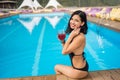 Portrait happy girl holding a cocktail, smiling and sitting on the edge of swimming pool at the resort Royalty Free Stock Photo