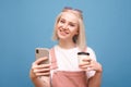 Portrait of a happy girl standing on a blue background with a smartphone and a cup of coffee in hand, looking into the camera and Royalty Free Stock Photo