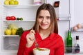 Portrait of happy glad young woman with attractive look dressed in red shirt, eats delicious vegetable salad, stands at kitchen ne Royalty Free Stock Photo