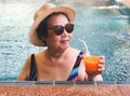 Happy and healthy Asian senior  woman wearing straw hat and sunglasses  drinking  orange juice  in the swimming pool, smiling and Royalty Free Stock Photo