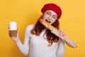 Portrait happy hungry woman holding paper bag with long white bread baguette and biting it, stands with coffee to go, wearing red Royalty Free Stock Photo