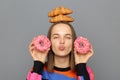 Portrait of happy joyful woman in sweater holding donuts in hands, standing with croissant on her head, looking at camera with