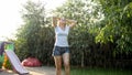 Portrait of happy laughing young woman with long hair in wet clothes dancing under warm rain in garden. Family playing Royalty Free Stock Photo